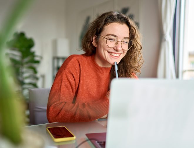 A younger woman in glasses holding a pen and smiling at her computer