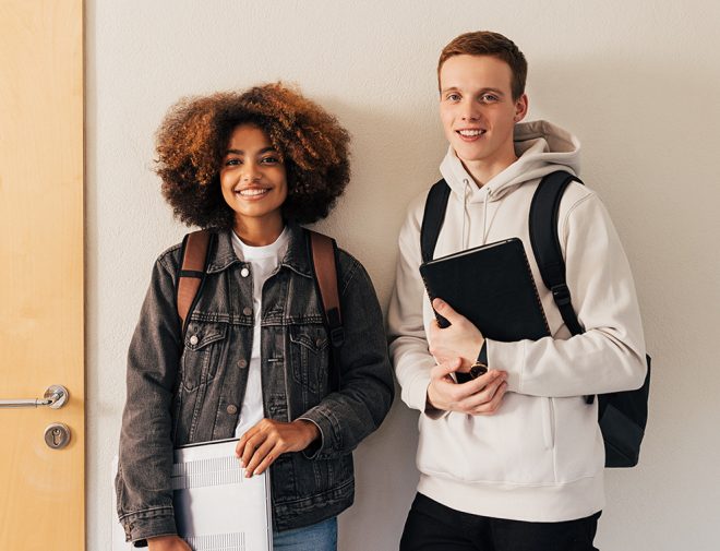 Two teen students standing against a wall holding books and a laptop