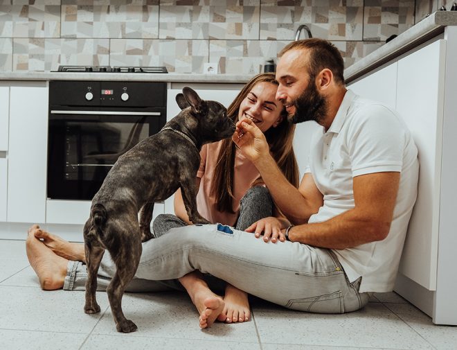 A couple sitting on the kitchen floor and feeding a treat to a small dog