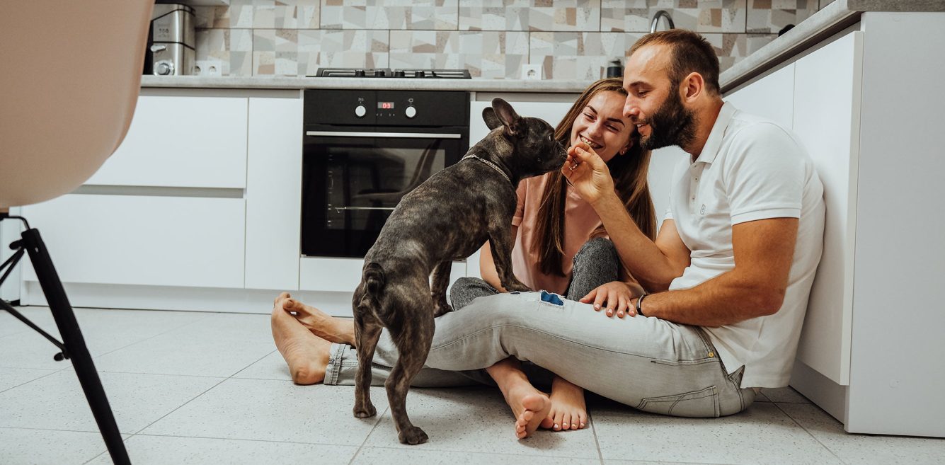 A couple sitting on the kitchen floor and feeding a treat to a small dog