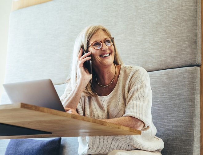 An older woman sitting in a booth with a laptop in front of her while she is on the phone and smiling