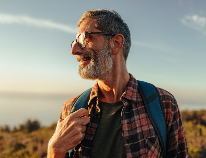 An older man with a beard and glasses hiking and smiling while looking towards the sun