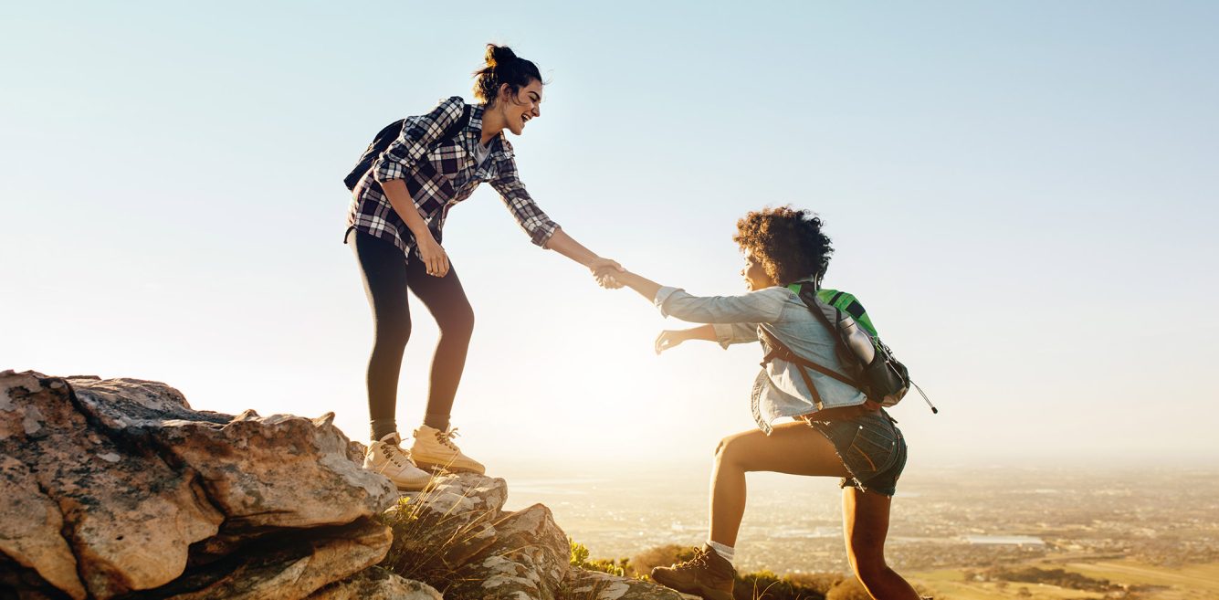 Two women hiking, one is helping the other on to a rock