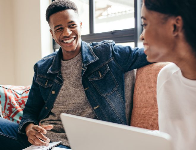 A young man and woman sitting on a couch smiling at each other, the man has a notepad in his lap and the woman has a laptop