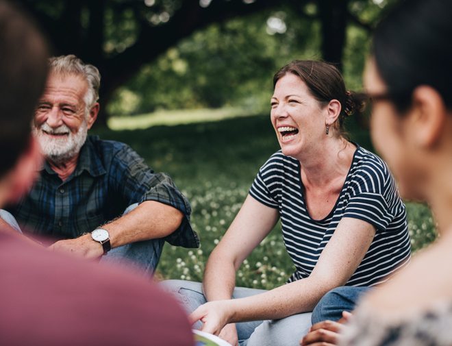 A middle aged woman sitting in a circle with other people laughing outdoors