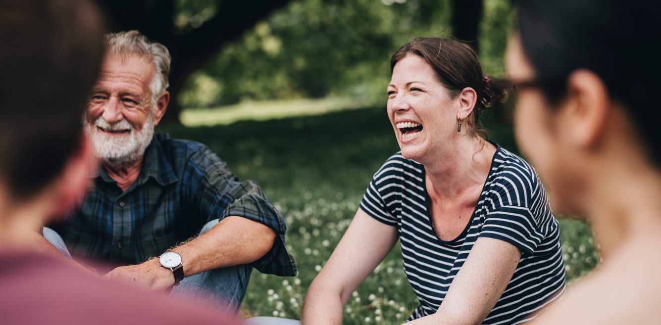 A middle aged woman sitting in a circle with other people laughing outdoors