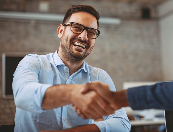 A man in glasses smiling and shaking hands with a person that is out of frame