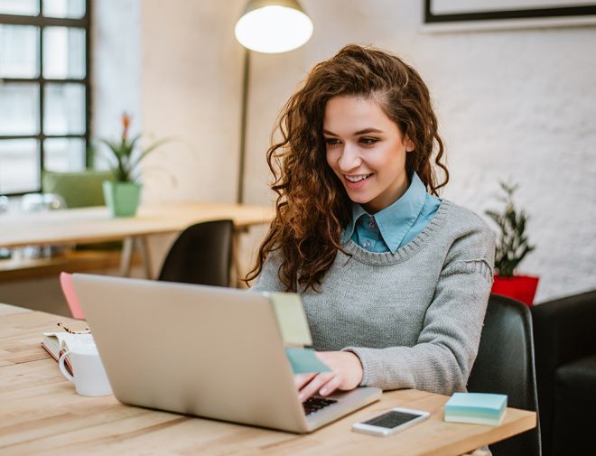 A woman with long curly hair smiling and working on a laptop