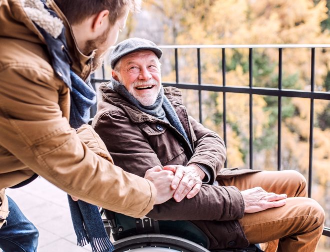 An older man in a wheelchair smiling and holding the hand of a younger man