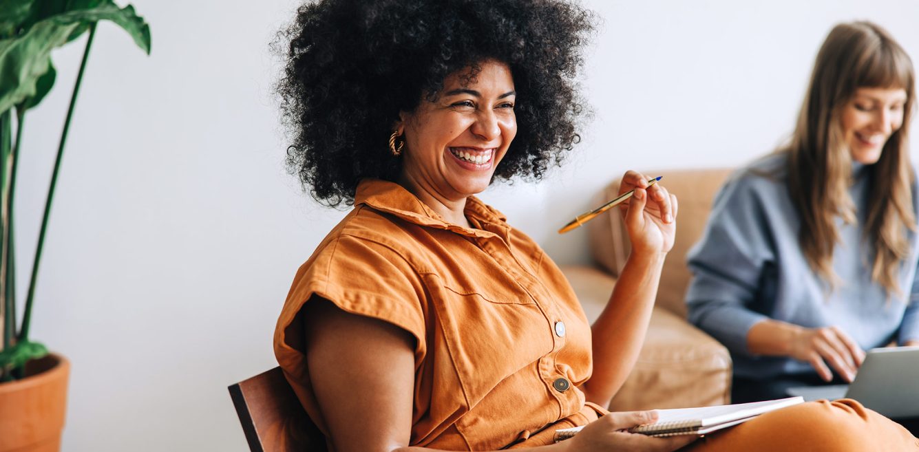An African American woman in an orange romper smiling and holding a pen