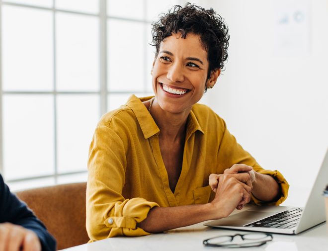 A woman with short curly hair sitting in front of a laptop and smiling at the person next to her